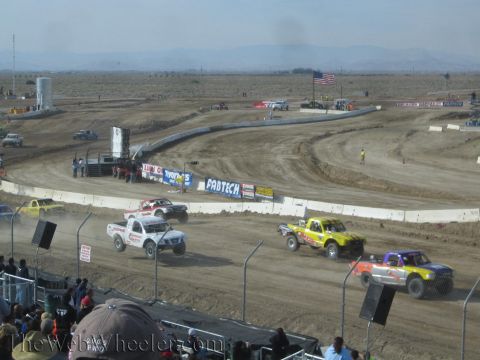 1990 Ford Bronco - white truck leads purple and yellow off-road race trucks around a dusty dirt oval lined with Fabtech banners under a hazy desert sky.