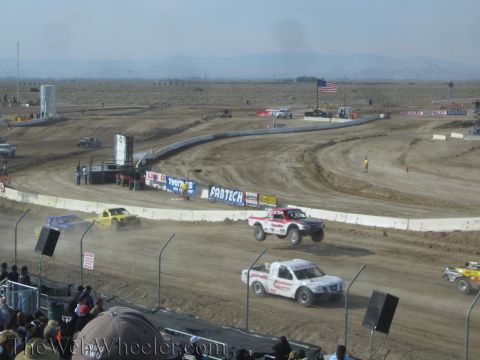 1990 Ford Bronco - white-and-red off-road race truck leads others around a dusty dirt oval lined with sponsor banners, seen from the grandstand.