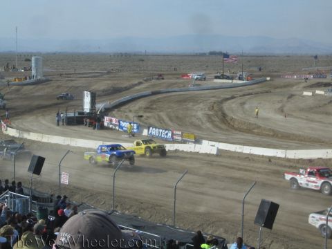1990 Ford Bronco - Blue and yellow off-road race trucks corner side by side on a dusty dirt oval lined with a Fabtech banner under a hazy desert sky.