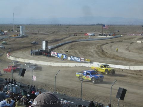 1990 Ford Bronco - Yellow and blue off-road race trucks run a dusty dirt oval lined with Fabtech and other sponsor banners, viewed from the grandstand.