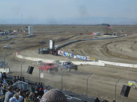 1990 Ford Bronco - Red and white off-road race trucks corner on a dusty dirt oval lined with Fabtech banners, with a desert valley and US flag beyond.
