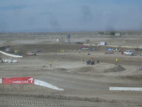 1990 Ford Bronco - wide view of a dusty dirt off-road course with scattered race trucks, track workers in vests, and a Kumho Tires banner in the foreground.