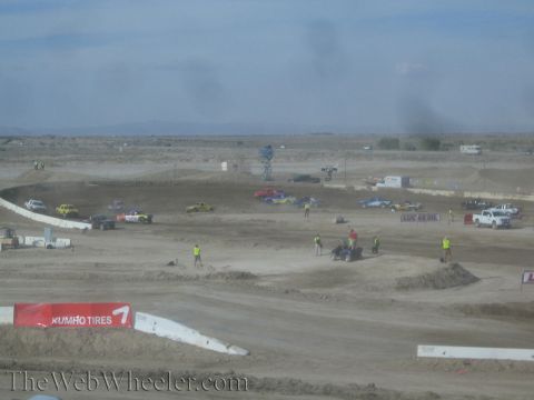 1990 Ford Bronco - Off-road race trucks and track crew spread across a sprawling dirt course in the desert, with a Kumho Tires banner along the near wall.