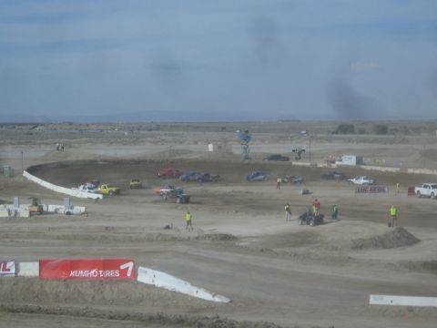 1990 Ford Bronco - Off-road race trucks and workers populate a sweeping dirt course with a tall light tower, jumps, and a Kumho Tires banner in the desert.