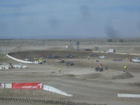1990 Ford Bronco - hazy wide view of a dirt off-road race track with several trucks, track crew, jumps, and a Kumho Tires banner in the desert.