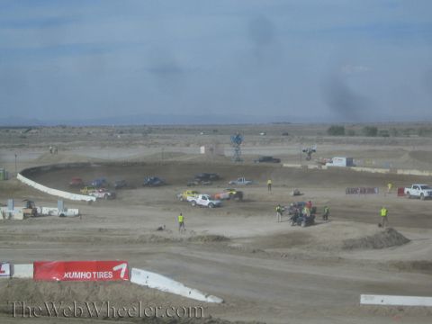 1990 Ford Bronco - Off-road race trucks and a cluster of track crew gather on a banked dirt course with jumps and a Kumho Tires banner in the desert.