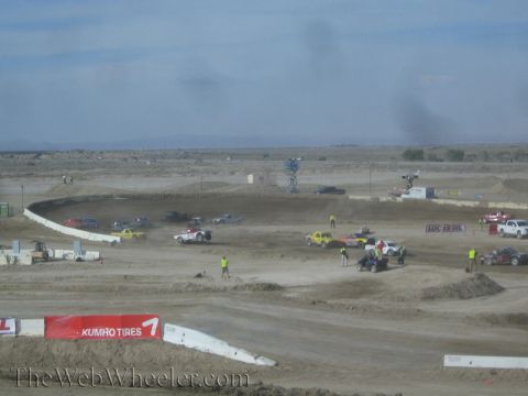 1990 Ford Bronco - Off-road race trucks navigate a dusty dirt course while crew in safety vests work the infield, with a Kumho Tires banner along the wall.