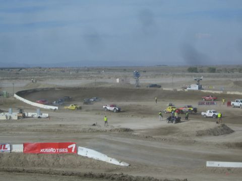 1990 Ford Bronco - Off-road race trucks and support pickups spread across a dusty dirt course with a light tower and Kumho Tires banner in the desert.