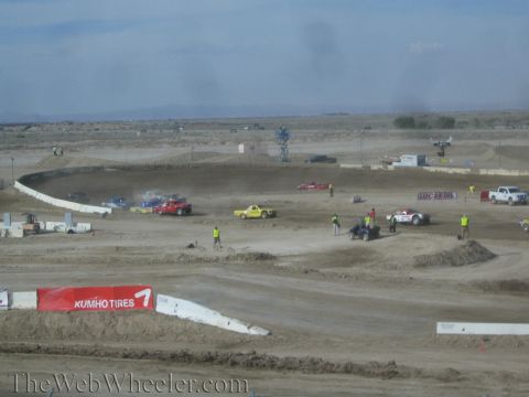 1990 Ford Bronco - Red and yellow off-road race trucks sit on a dusty dirt course as crew in vests work the infield near a Kumho-style banner in the desert.