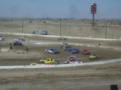 1990 Ford Bronco - cluster of colorful off-road race trucks groups up on a dusty dirt oval beneath a tall roadside sign, with a blue lift in the background.