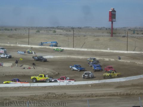 1990 Ford Bronco - Numerous colorful off-road race trucks bunch together on a dusty dirt oval near jumps and a tall roadside sign in the desert.
