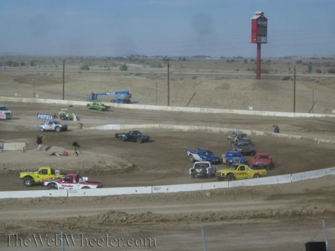 1990 Ford Bronco - field of colorful off-road race trucks spreads across a dusty dirt oval with jumps, a blue lift, and a tall roadside sign behind.