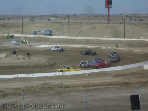 1990 Ford Bronco - Off-road race trucks group on a banked dusty dirt oval with jumps and a blue boom lift, beneath a tall red roadside sign.
