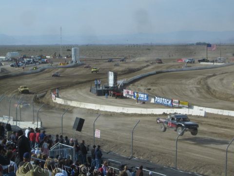 1990 Ford Bronco - off-road race truck runs a dusty dirt oval lined with Fabtech and other sponsor banners and a US flag, with spectators watching nearby.