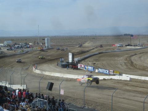 1990 Ford Bronco - orange short-course off-road race truck catches air on a desert dirt oval with US flag and sponsor banners in the background.