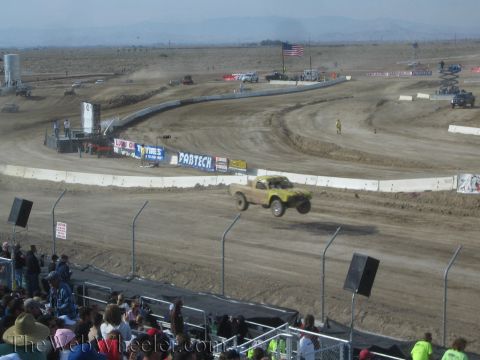 1990 Ford Bronco - yellow off-road race truck flies off a jump on a dusty desert dirt track lined with sponsor banners and fencing.