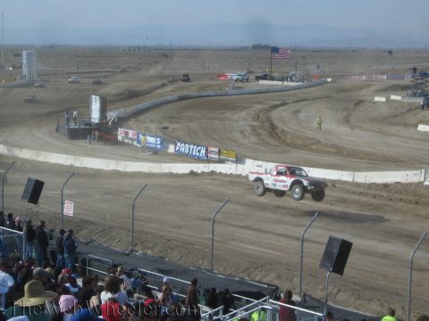 1990 Ford Bronco - white and orange off-road race truck launches off a dirt jump at a desert short-course track with mountains behind.