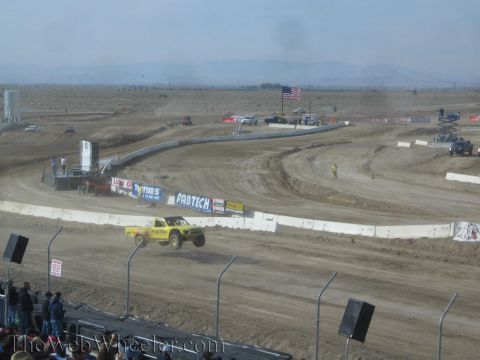 1990 Ford Bronco - yellow off-road race truck rounds a banked turn on a dusty desert dirt track in front of grandstand fencing.