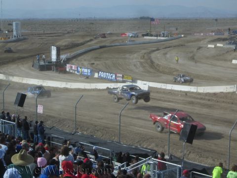 1990 Ford Bronco - Blue and red off-road race trucks run close together on a banked desert dirt oval as spectators watch from the stands.