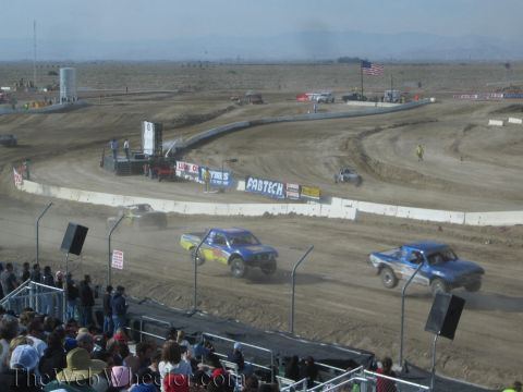 1990 Ford Bronco - yellow and a blue off-road race truck run side by side through a turn on a desert dirt track lined with banners.