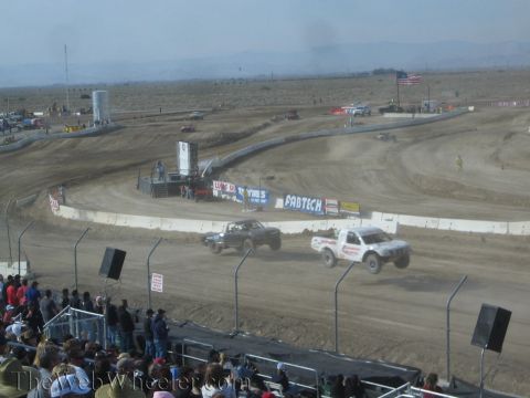 1990 Ford Bronco - Two off-road race trucks throw up dust as they corner on a desert dirt track with a US flag and banners behind.