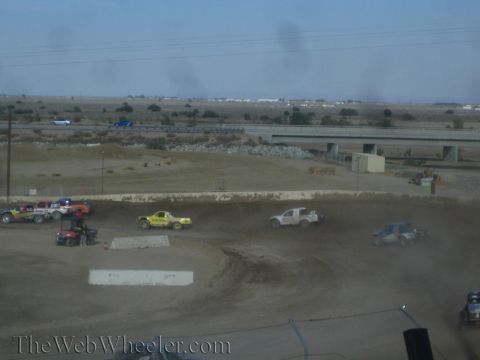 1990 Ford Bronco - Several off-road race trucks and an ATV gather on a dirt track with a freeway overpass and desert in the background.