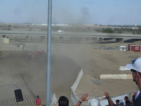 1990 Ford Bronco - red off-road race truck lies rolled on its side in a dusty turn as spectators in the foreground watch and clap.