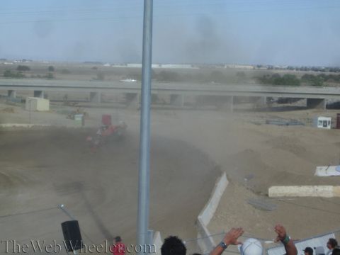 1990 Ford Bronco - red off-road race truck sits overturned in a dust cloud on a dirt track as spectators raise their hands in the foreground.