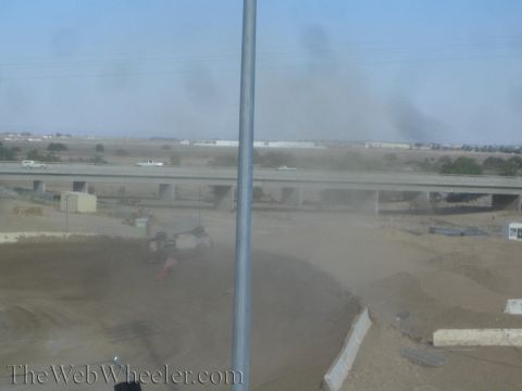 1990 Ford Bronco - flipped off-road race truck rests on a dirt track with a freeway overpass and warehouses in the hazy distance.