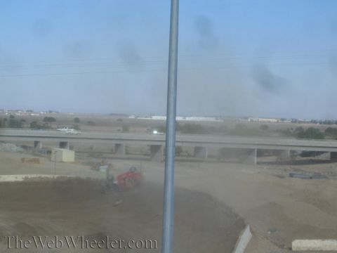 1990 Ford Bronco - red off-road race truck sits crashed in a dusty dirt turn with a freeway overpass and desert buildings behind.