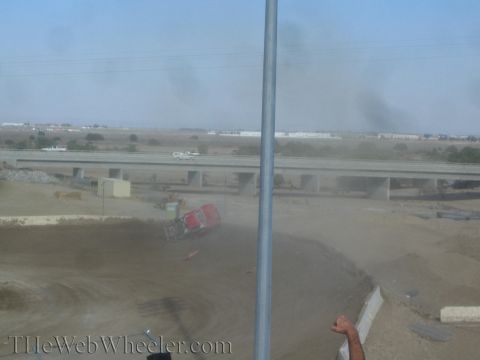 1990 Ford Bronco - red and yellow off-road race truck lies on its side in a dust cloud on a dirt track near a freeway overpass.