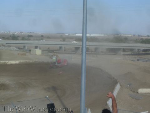 1990 Ford Bronco - red off-road race truck sits askew in a dusty turn of a dirt track with a freeway overpass in the background.