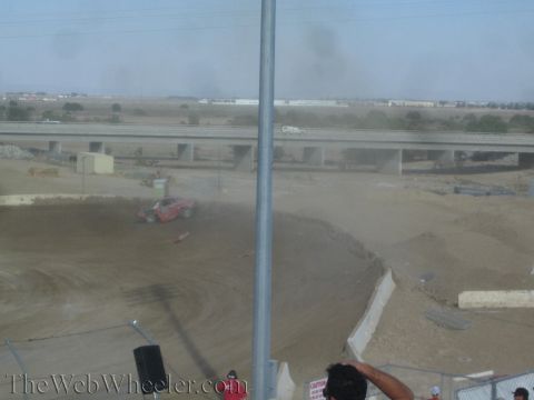 1990 Ford Bronco - red off-road race truck moves through a dusty dirt turn as a freeway overpass and desert stretch behind it.