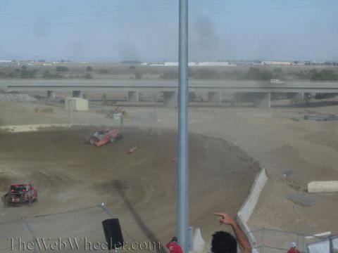 1990 Ford Bronco - Two red off-road race trucks run through a dusty dirt turn with a freeway overpass and warehouses behind them.
