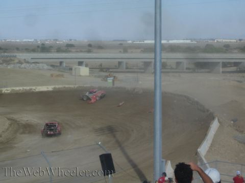 1990 Ford Bronco - Red off-road race trucks race through a banked dirt turn kicking up dust, with a freeway overpass in the background.