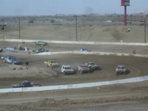 1990 Ford Bronco - field of colorful off-road race trucks lines up and races on a desert dirt oval with power lines and a road behind.