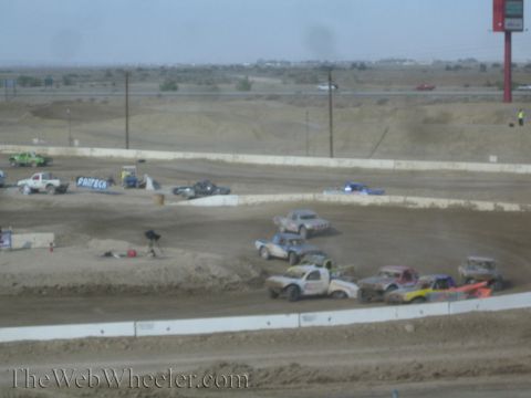 1990 Ford Bronco - Off-road race trucks stage and run on a dusty desert dirt oval with sponsor banners and a flat desert horizon behind.