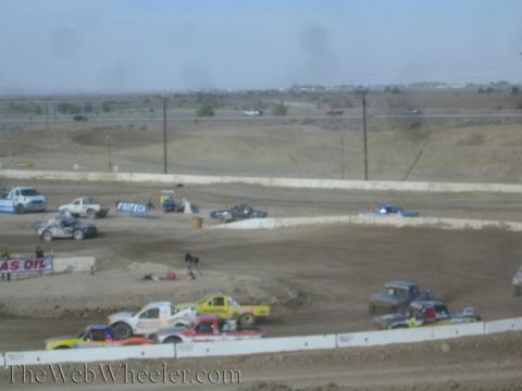 1990 Ford Bronco - pack of colorful off-road race trucks groups together on a desert dirt oval with a highway and power lines behind.