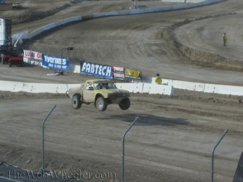 1990 Ford Bronco - tan off-road race truck rounds a turn on a desert dirt track past Fabtech and other sponsor banners along the wall.