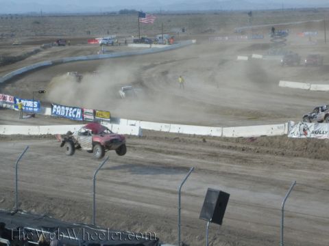 1990 Ford Bronco - red off-road race truck rounds a turn while another truck throws up dust on a desert dirt track with banners and a US flag.