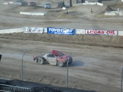 1990 Ford Bronco - red off-road race truck runs along a dirt track wall lined with Toyo Tires, Lucas Oil and Bully Dog banners.