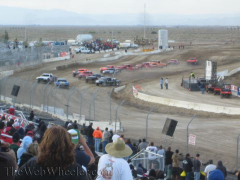 1990 Ford Bronco - Spectators in the grandstand watch a field of off-road race trucks staged on a desert dirt oval at dusk.