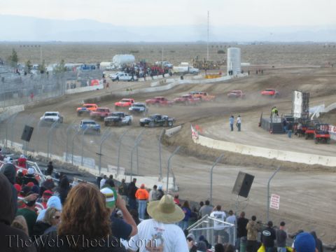 1990 Ford Bronco - Spectators watch a pack of off-road race trucks navigate a dusty desert dirt short-course oval.