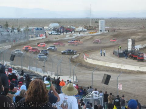 1990 Ford Bronco - line of off-road race trucks rounds a turn on a desert dirt track as a crowd looks on through the catch fence.