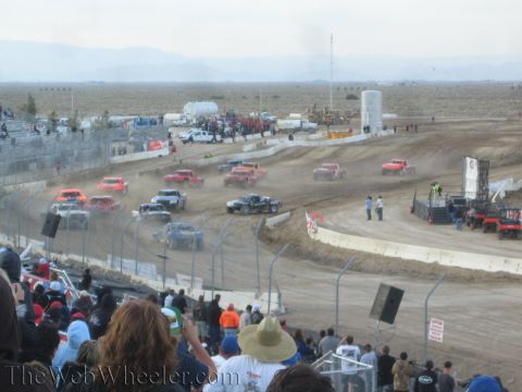 1990 Ford Bronco - Off-road race trucks spread across a dusty desert dirt course with mountains in the background.