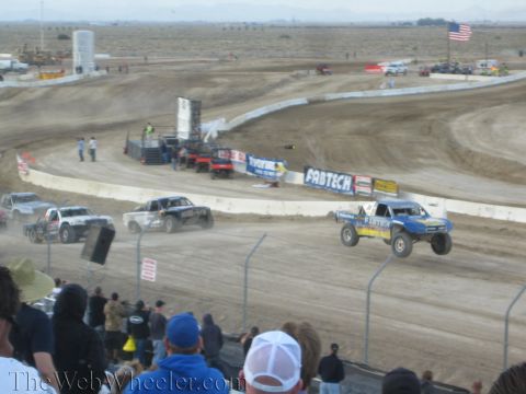 1990 Ford Bronco - blue Fabtech-liveried off-road race truck leads rivals on a desert dirt track beneath a US flag.