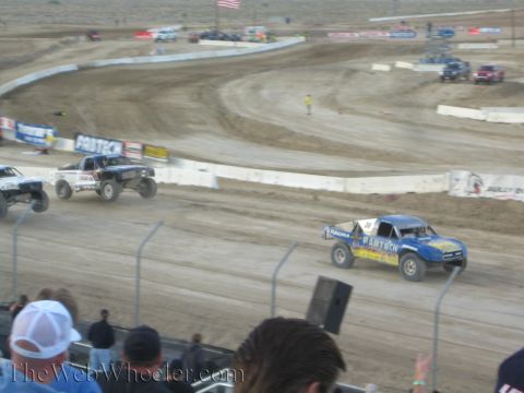 1990 Ford Bronco - blue Fabtech off-road truck and a white truck race on a banked desert dirt course behind a fence.