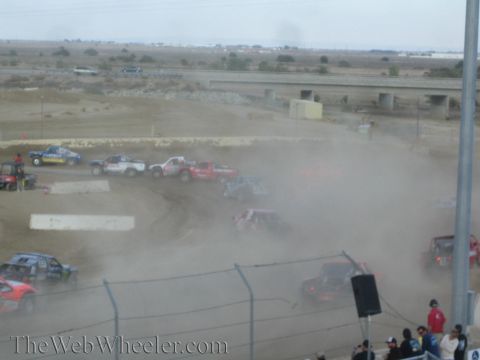 1990 Ford Bronco - Off-road race trucks churn up heavy dust on a dirt course with a highway overpass behind.