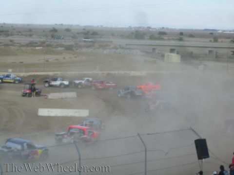 1990 Ford Bronco - cluster of off-road race trucks raises dust on a desert dirt course near a highway overpass.