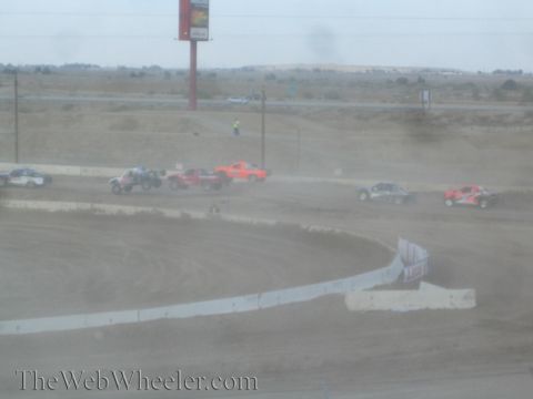 1990 Ford Bronco - Several off-road race trucks spread across a desert dirt course below a red billboard.